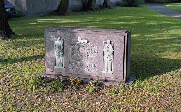 Ein Mahnstein mit Bildern und Text, umgeben von Gras, einer Straße, Bäumen, einem geparkten Auto und einer Wand im Hintergrund.