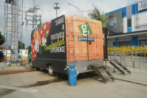Ein Lastwagen vor einem Gebäude mit einer Tafel, auf der "Teilen Sie ein positiv uberladenes Erlebnis" steht, mit Stufen, einem Zylinder auf dem Boden, Geländern, elektrischen Masten mit Drähten, Laternenmasten, Bäumen und einem bewölktem Himmel im Hintergrund.