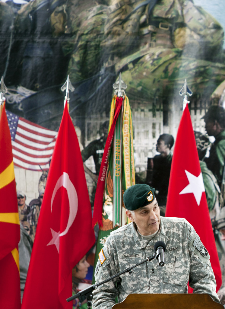 Ein Soldat steht auf einem Podium mit einem Mikrofon, mit verschiedenen Flaggen und einem Plakat von Militärpersonal im Hintergrund.