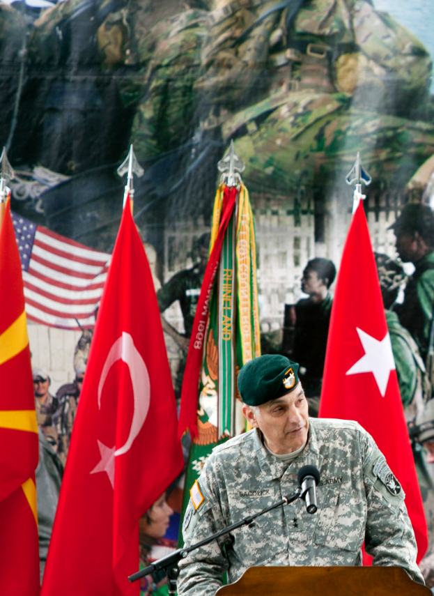 Ein Soldat steht auf einem Podium mit einem Mikrofon, mit verschiedenen Flaggen und einem Plakat von Militärpersonal im Hintergrund.