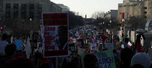 Menschen protestieren auf einer Straße und halten Plakate, mit Gebäuden, Bäumen, Laternen und Himmel im Hintergrund.