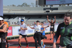 Gruppe von Menschen, die auf einer Outdoor-Sportanlage laufen, mit Bannern, einem Display, einem Gebäude, Bäumen und einer bewölkten Himmel; einige Läufer tragen Turnschuhe und Hüte.
