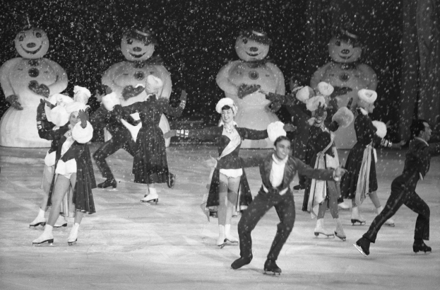 Eine Gruppe von Menschen in Kostümen und Schlittschuhen fährt auf einem Eisplatz im Schnee, mit Schneemännchen im Hintergrund, in Schwarz-Weiß aufgenommen.