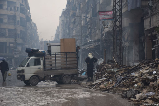 Ein Lastwagen fährt auf einer Straße an einem Schutthaufen vorbei, während Menschen auf der Straße gehen, einer davon hält einen Schirm, und Gebäude, Pfosten und Schilder mit Text im Hintergrund unter einem klaren Himmel.