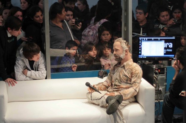 Eine Gruppe von Menschen, die ein Puppenspiel fotografieren, bei dem eine Person einen Bildschirm ansieht und eine puppenähnliche Person auf einem Sofa sitzt und ein Mikrofon hält.