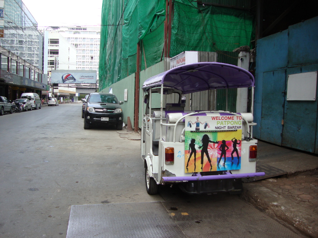 Gebäude mit parkenden Autos, eine Werbetafel, ein bewölkter Himmel und ein Auto-Rikscha auf der Seite.