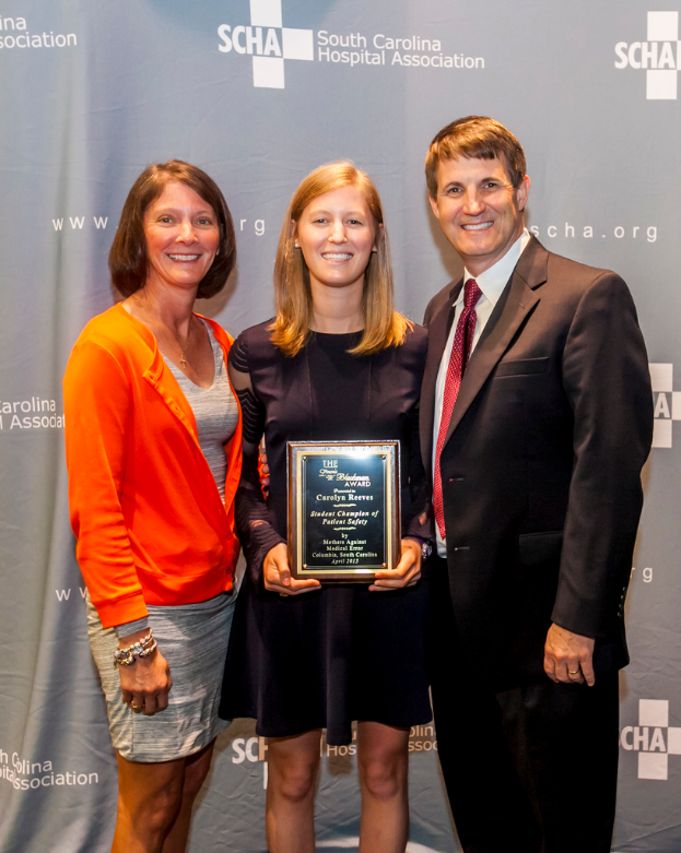 Drei Menschen, zwei Frauen und ein Mann, stehen zusammen. Der Mann trÃ¤gt einen schwarzen Anzug, eine Frau ist in einem orange-schwarzen Kleid, die andere Frau in einem schwarzen Kleid mit einer TrophÃ¤e in der Hand. Sie stehen vor einem grauen Hintergrund.