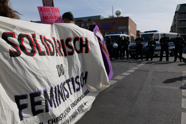 Gruppe von Menschen, die eine Straße entlanggehen und eine 'Solidarität und Feminismus'-Schleife halten, mit geparkten Fahrzeugen, Gebäuden, einer Schüsselantenne und einem klaren blauen Himmel im Hintergrund.