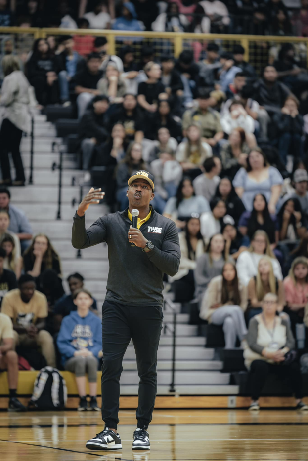 Ein Mann mit M├╝tze h├Ąlt ein Mikrofon auf einem Basketballfeld, mit einer Gruppe von Menschen auf einer Treppe und Taschen auf dem Boden hinter ihm.