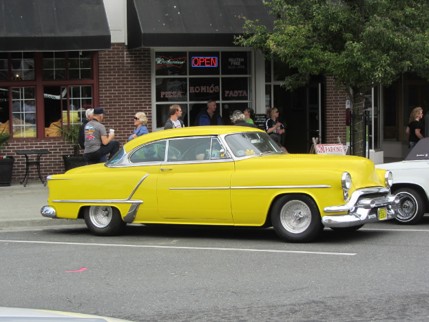 Eine Straßenszene mit Autos, einigen Fußgängern, Stühlen, einem Baum und einem Gebäude im Hintergrund.