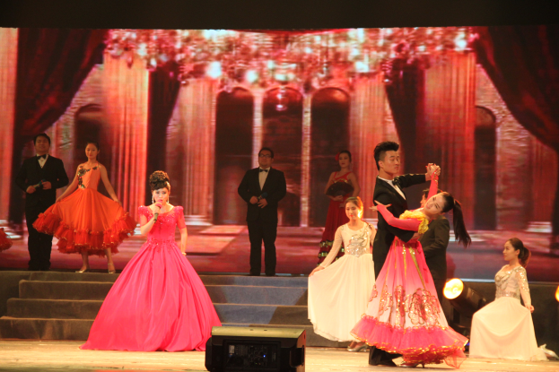 Eine Gruppe von Menschen steht auf einer Bühne mit Stufen und einem Bildschirm im Hintergrund, beleuchtet von Scheinwerfern, mit einem Lautsprecher unten, was eine Tanzvorstellung vermuten lässt.