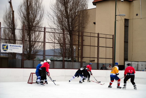 Menschen beim Eishockey auf einer Eisbahn mit GebÃ¤uden, BÃ¤umen, einer StraÃŸenlaterne, einem Namensschild und ZÃ¤unen im Hintergrund bei einem klaren Himmel.