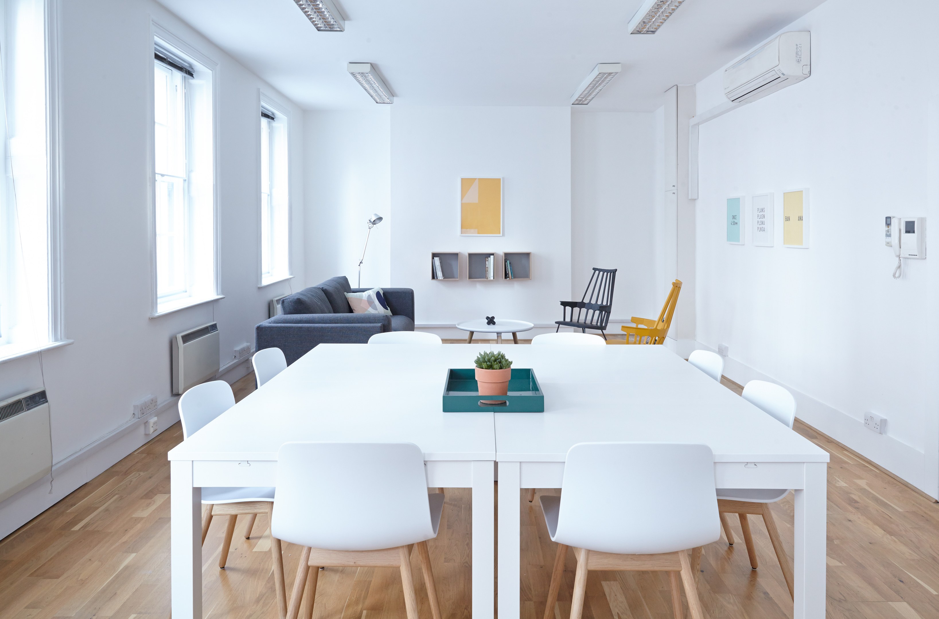 Ein weißer Tisch mit einer Topfpflanze in einer Schale, umgeben von Stühlen, einem Sofa mit Kissen, einer Lampe, Regalen mit Gegenständen, einer Klimaanlage, Fotorahmen an der Wand, Fenstern und Deckenleuchten in einer Wohnungseinrichtung.