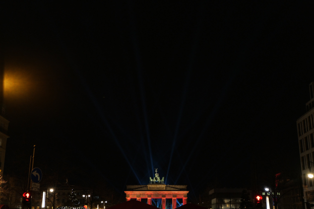 Brandenburger Tor in Berlin bei Nacht, beleuchtet von Straßenlaternen, mit Gebäuden, Bäumen, Polen, Verkehrszeichen, Schildern und Zelten im Vordergrund und einem dunklen Himmel im Hintergrund.