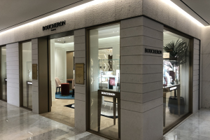 Eintritt in ein Boutique in einem Einkaufszentrum mit Glaswänden und -türen, beleuchteter Decke, Möbeln wie Stühlen und Tischen, einer Pflanze und sichtbarem Boden.