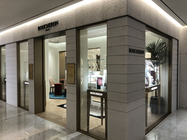 Eintritt in ein Boutique in einem Einkaufszentrum mit Glaswänden und -türen, beleuchteter Decke, Möbeln wie Stühlen und Tischen, einer Pflanze und sichtbarem Boden.