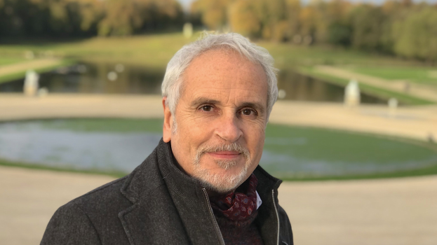 Ein Mann in einem weißen Hemd steht neben einem Teich in einem Park, umgeben von Bäumen und Gras unter einem klaren blauen Himmel, mit einem ernsten Gesichtsausdruck.