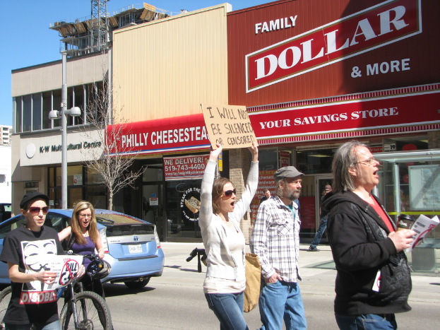 Eine Gruppe von Menschen demonstriert auf einer Straße mit Gebäuden, einem Mast und einem Baum im Hintergrund, einem Auto und einem Fahrrad auf der linken Seite und dem Himmel oben