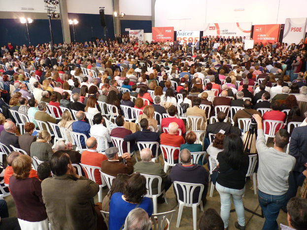 Ein volles Stadion mit vielen sitzenden und stehenden Menschen, eine Bühne mit zwei Rednern an einem Podium und zahlreiche Banner und Lichter.