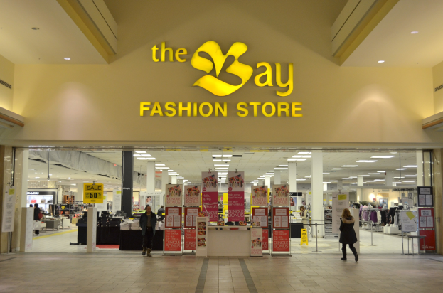 Außenansicht eines Bay Fashion Stores in einem Einkaufszentrum mit Menschen, Texttafeln an Wänden, Säulen, Deckenleuchten und Hintergrundobjekten.