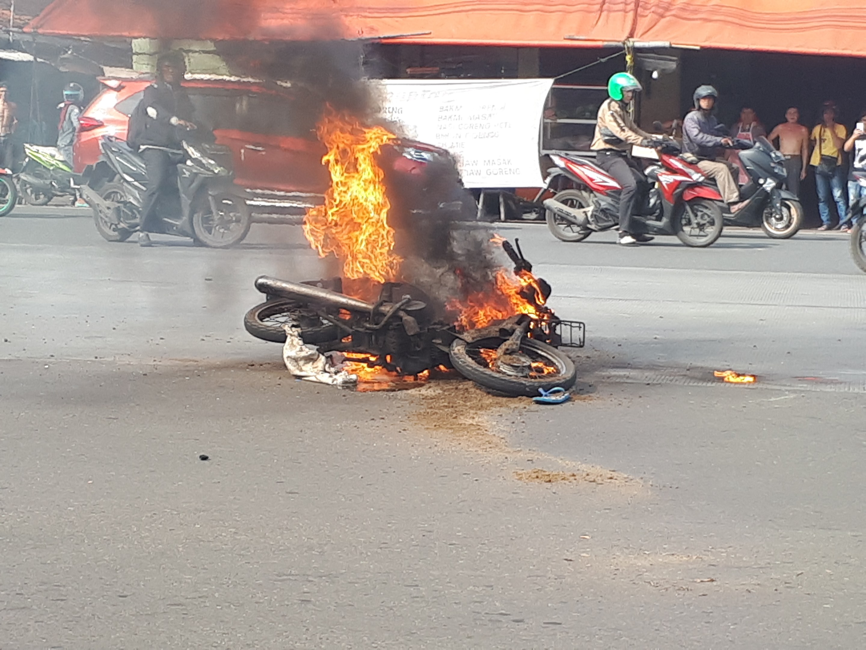 Motorrad in Flammen auf der Straße mit einem Auto und einem Van im Hintergrund, umgeben von Menschen, einige mit Helmen, mit einer Fahne und einem Zelt im Bild.
