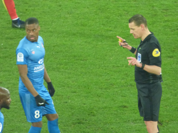 Ein Schiedsrichter spricht mit einem Füßballspieler auf dem Feld, umgeben von einer Gruppe von Menschen, wobei eine Person ein Objekt hält und die Beine einer anderen Person im Hintergrund zu sehen sind.