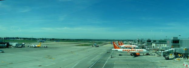 Eine Flughafenszene mit Flugzeugen, Gebäuden, Polen, Fahrzeugen und Bäumen unter einem Himmel mit einer Straße unten und einem Zaun im Vordergrund.