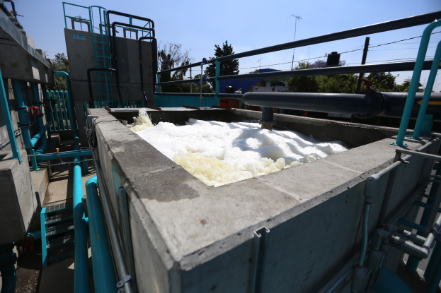 Ein großer Tank mit weißem Schaum steht auf einer Betonwand, umgeben von Metallstangen, Treppen, Geländern und anderen Gegenständen, mit Bäumen, Pfählen, Drähten und einem klaren blauen Himmel im Hintergrund.