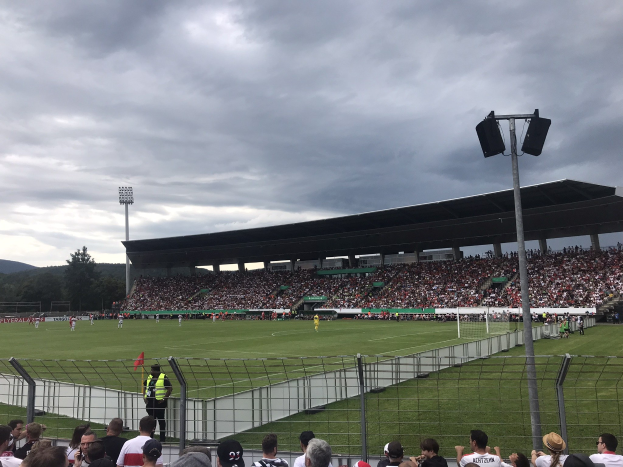 Große Zuschauermenge bei einem Fußballspiel in einem eingezäunten Stadion unter künstlicher Beleuchtung, mit Bäumen und Hügeln im Hintergrund unter einem bewölkten Himmel.