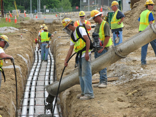 Eine Baustelle mit Arbeitern in Jacken und Helmen, Absperrbaken, Zaun, Bäume, Pfosten, Erde und Rohrleitungen im Hintergrund.