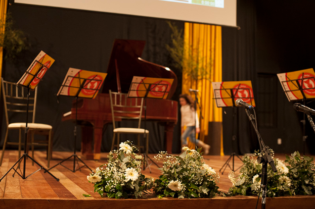 Eine Bühne mit Mikrofonen auf Ständern, Blumenbouquets, Stühlen, Notenständern mit Noten, einem Flügel, einer Person, die läuft, und Bäumen, Vorhängen und einer Projektionswand im Hintergrund.