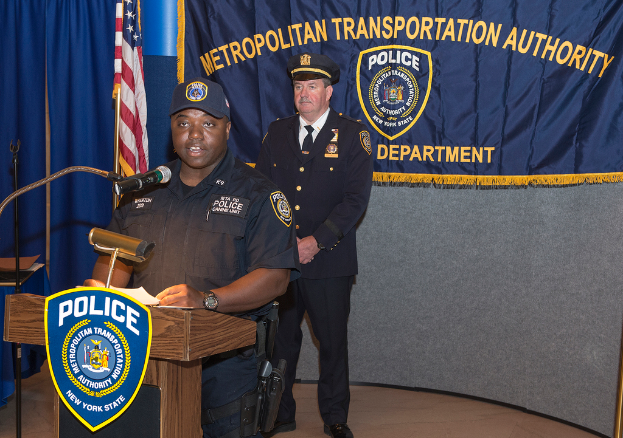 Zwei Polizisten stehen vor einem Rednerpult, einer hält Papiere und spricht in ein Mikrofon, mit einer Fahne, einem Tisch und einer Tafel mit Symbolen und Text in der Nähe.