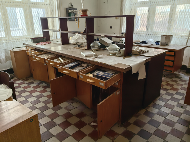 Eine alte Küche mit kariertem Boden und hölzernen Schänken, mit einem Tisch mit verschiedenen Gegenständen und einem Bett mit einem Kissen auf der linken Seite, mit Fenstern und Vorhängen im Hintergrund, die eine verlassene Krankenhausatmosphäre vermuten lassen.