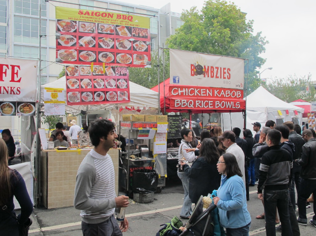 Menschen stehen auf einer Straße außerhalb der Stadt, mit einem Mann in einem grauen und weißen T-Shirt, der eine Wasserflasche hält und eine Frau in einer blauen Jacke, die einen Kinderwagen schiebt. Hinter ihnen sehen wir Stände mit weißen und roten Zelten, farbige Banner und Gebäude, Bäume und eine Straßenlaterne im Hintergrund.