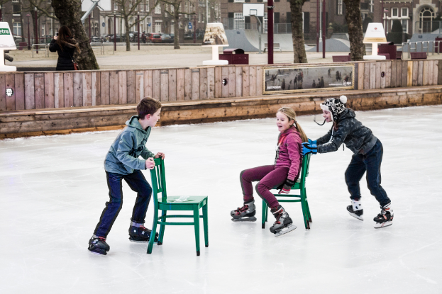 Kinder beim Skifahren vor einem Spielplatz, mit drei Kindern und zwei St├╝hlen in der Mitte und Geb├Ąuden, B├Ąumen, B├Ąnken, Pfosten und einem Basketballfeld im Hintergrund.