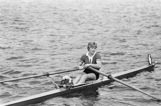 Eine Frau in einem weißen Hemd und blauen Shorts rudert ein Einerboot mit weißem Rumpf und blauem Segel auf einem sonnigen Gewässer, wobei sie zwei Paddel verwendet und ihr Haar zu einem Pferdeschwanz gebunden hat.