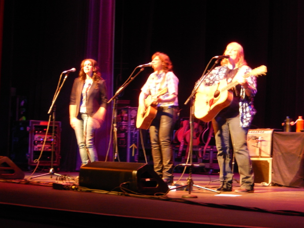 Drei Frauen performen auf der Bühne eines Musikkonzerts, singen jeweils in ein Mikrofon und spielen eine Gitarre, mit einem Tisch voller Wasserflaschen und zusätzlichen Gitarren dahinter.