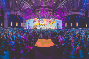 Eine große Menschenmenge, die auf Stühlen sitzt und einer hell erleuchteten Bühne mit mehreren stehenden Individuen zugewandt ist, wahrscheinlich bei einem Online-Marketing-Rockstars-Event, umgeben von Bildschirmen.