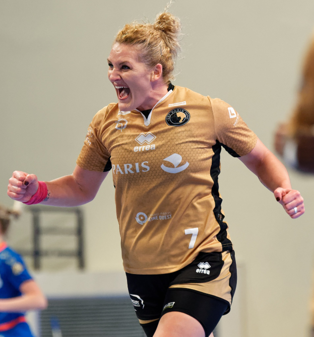 Eine Frau in braunem T-Shirt und schwarzen Shorts rennt auf einem Futsalplatz, feiernd mit triumphaler Miene, während zwei andere Frauen im Hintergrund sichtbar sind.