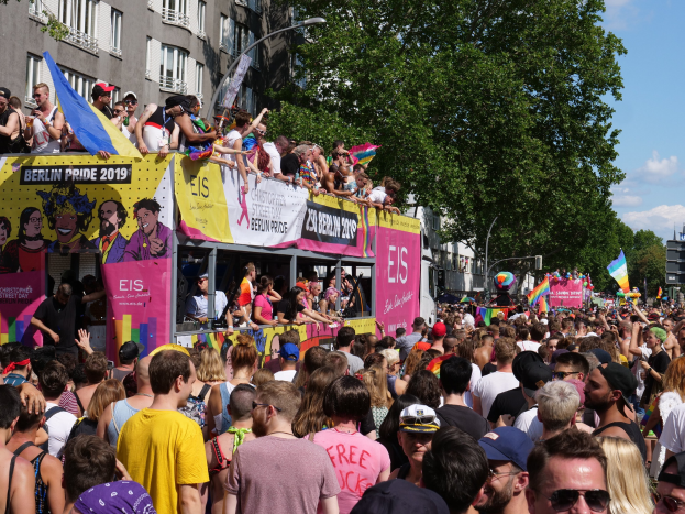 Eine große Menschenmenge marschiert auf der Straße während der Christopher Street Day Parade 2019 in Berlin, viele tragen Mützen und Goggles, halten Fahnen, mit Bannern und Gebäuden im Hintergrund unter einem bewölkten Himmel.