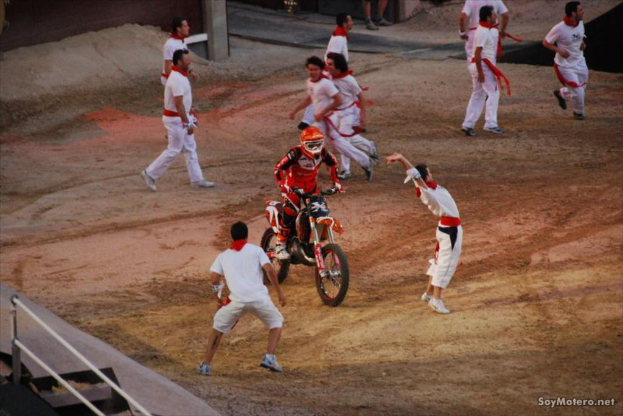 Eine Person in rotem Kleid und Helm fährt ein Motorrad in der Mitte eines Feldes, umgeben von Jungen in weißen Kleidern, die laufen.