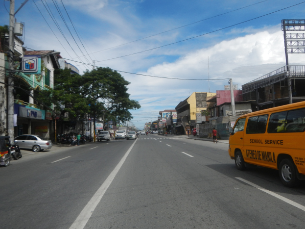 Ein gelber Schulbus fährt eine Straße entlang, die von hohen Gebäuden gesäumt ist, Menschen auf dem Bürgersteig, Bäumen, Strommasten mit Drähten und Schildern, unter einem bewölkten Himmel.