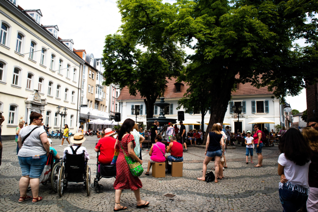 Eine Gruppe von Menschen, einige im Rollstuhl, geht eine Kopfsteinpflasterstraße in der Altstadt von Heidelberg entlang, mit Bäumen, Gebäuden, Laternen und einer Statue im Hintergrund bei einem bewölkten Himmel.