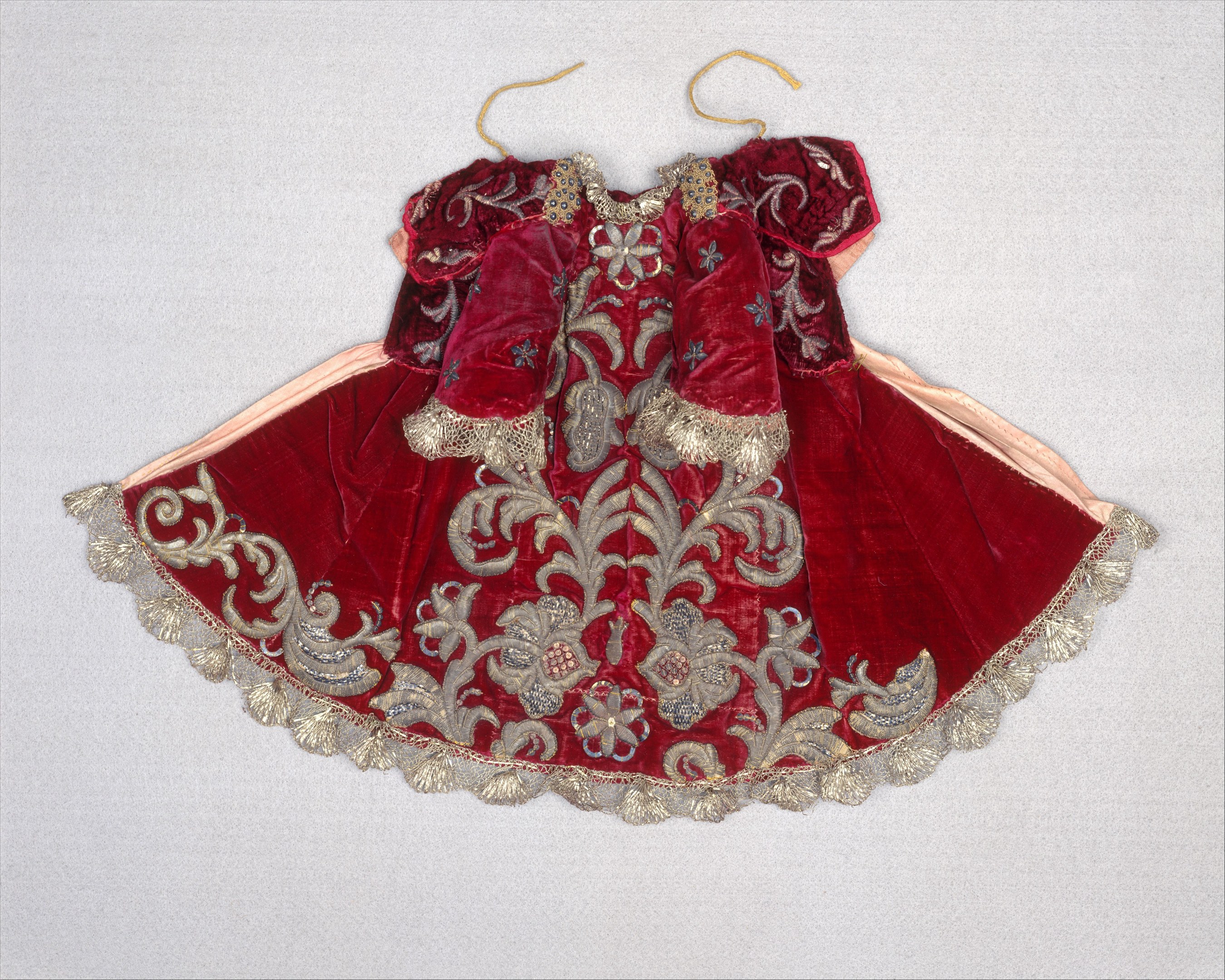 Ein rotes Samtkleid mit goldener Stickerei und detaillierten Designs, das an einem Bügel gegen einen weißen Hintergrund gehangen ist.