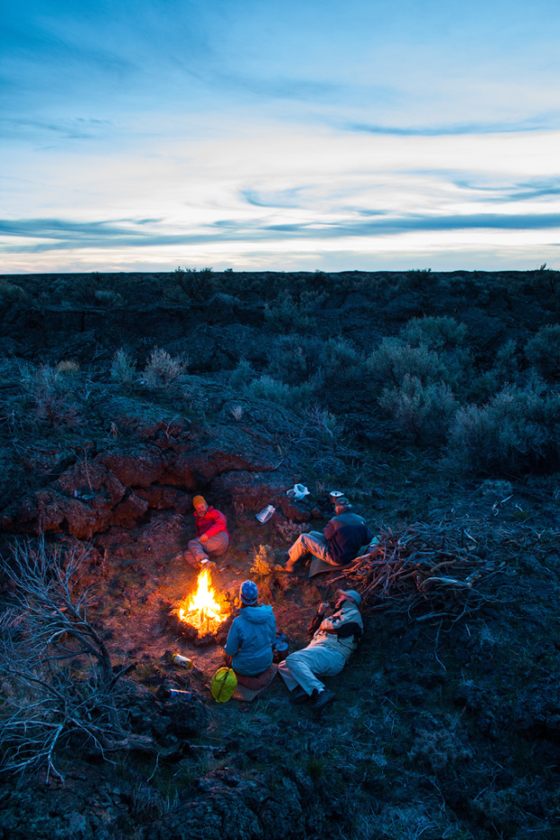 Ein Lagerfeuer mit Menschen, die darauf sitzen und liegen, mit Mützen tragend, einem Beutel und anderen Gegenständen auf dem Boden, umgeben von Bäumen und Himmel.