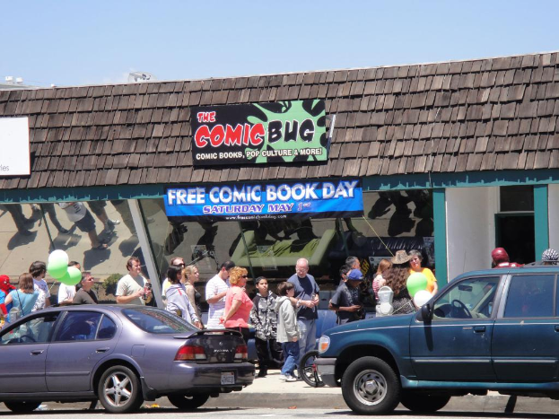 Gruppen von Menschen stehen vor einem Gebäude mit Glastüren und einem Dach, zwei Autos auf der Straße, grüne Luftballons, eine Fahne und ein Namensschild auf dem Dach, unter einem klaren Himmel.