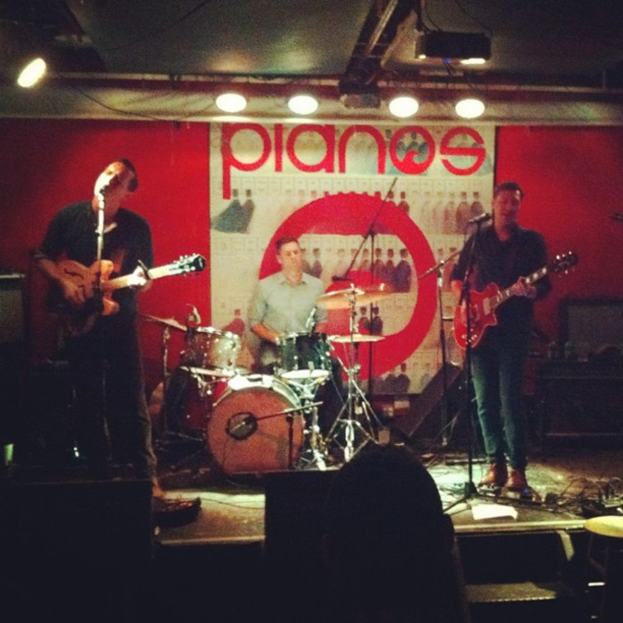 Drei Männer auf der Bühne: Einer spielt am Drumset in der Mitte, zwei spielen Gitarre und singen auf der linken und rechten Seite, mit einem Banner an der Rückwand und Lampen an der Decke.