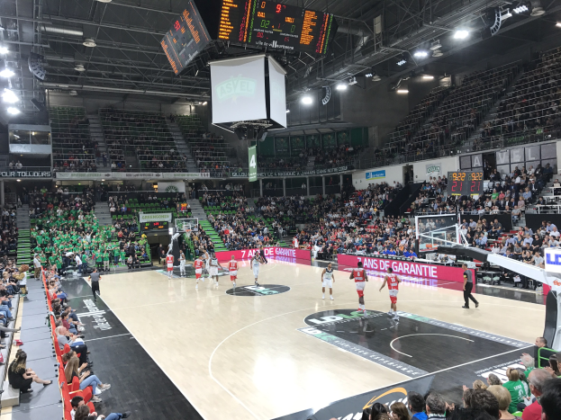 Basketballspiel in einer großen Halle mit Zuschauern, Anzeigetafeln mit "Asvel vs Galatasaray" und Deckenbeleuchtung und -bildschirmen.