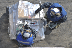 Zwei Männer in blauen Uniformen und Helmen arbeiten an einem großen Drohnenmodell, jeder hält Werkzeuge, mit einer Plastikabdeckung auf dem Boden.