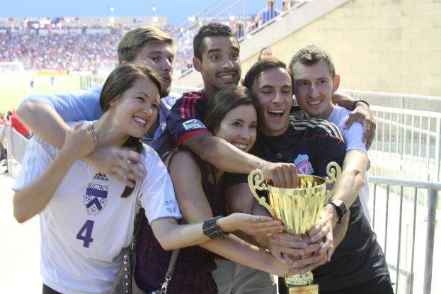Sechs Personen, darunter zwei Frauen und vier Männer, halten einen Pokal und lächeln, posieren für ein Foto vor einem Eisengeländer, mit einer Wand und einem Geländer oben rechts, Menschen und Lichter im Hintergrund, möglicherweise in einem Stadion.